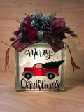 A lighted glass block with a vintage Christmas tree delivery truck design, accompanied by a festive arrangement on top.