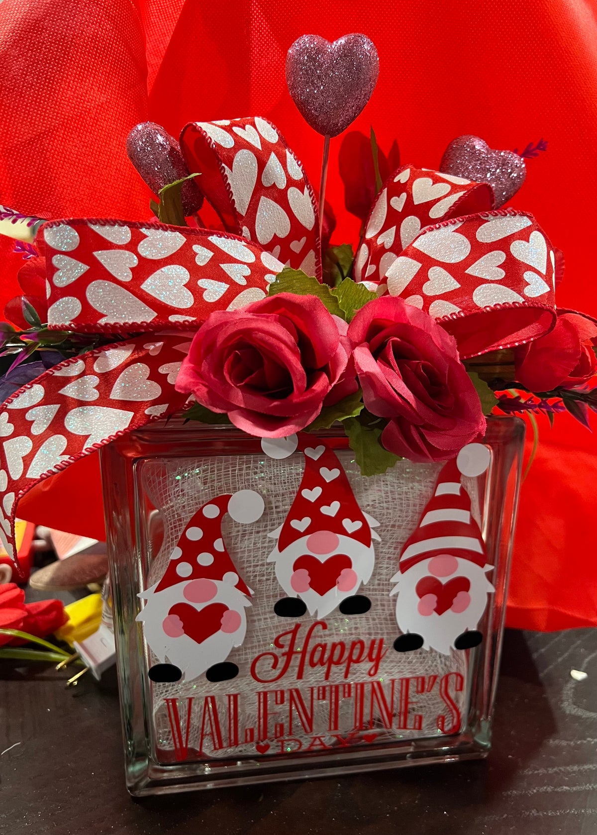 A Valentine's Day themed light box featuring a glass block adorned with red fabric, artificial red hearts, a pink rose, and gnomes wearing red and white hats.