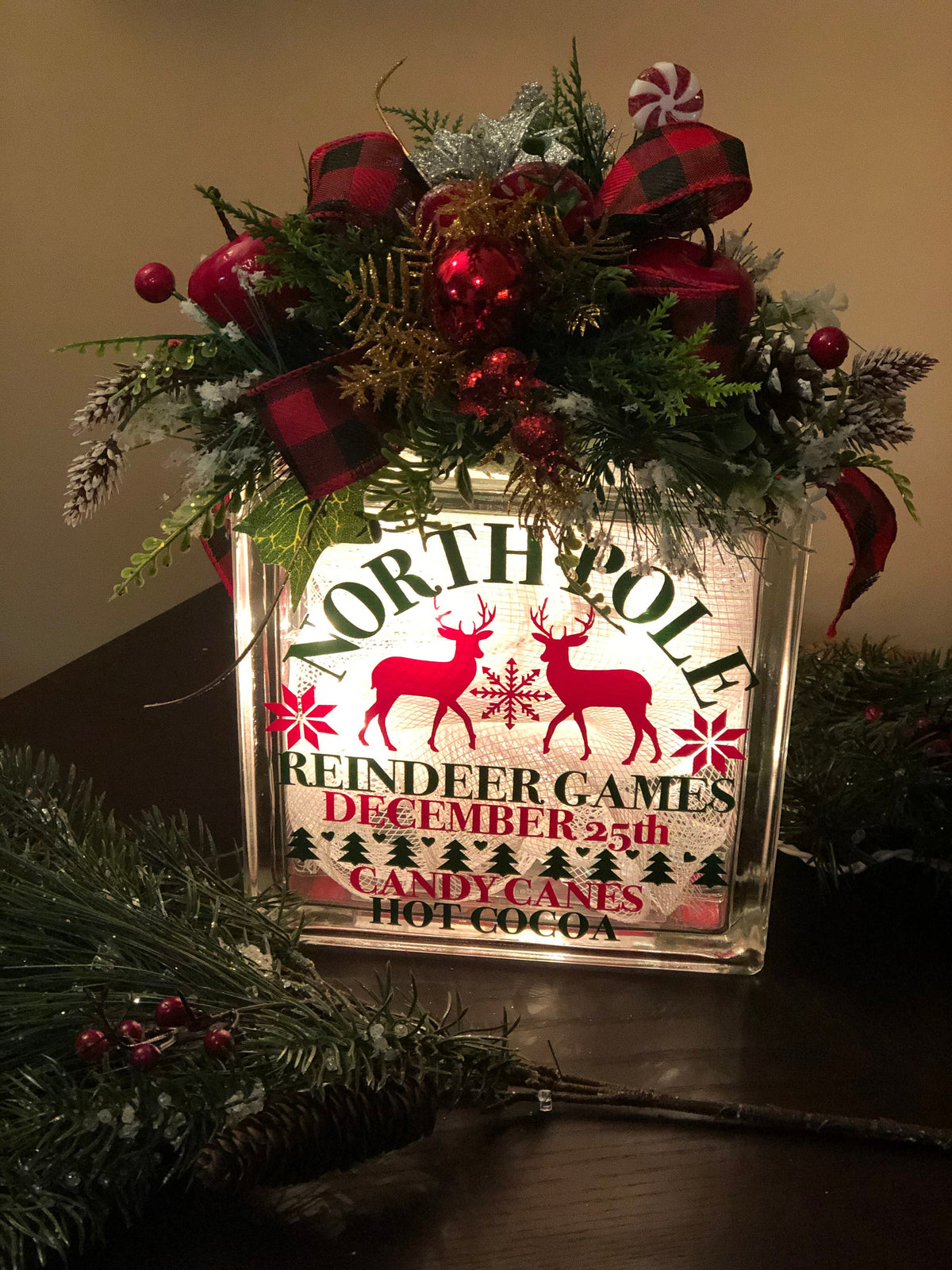 A decorative glass light box with 'Reindeer Games, North Pole, December 25th, Candy & Hot Cocoa' printed on it, adorned with a Christmas arrangement including red and green decorations and sitting on a table with evergreen branches.